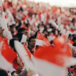 A crowd of Singaporeans cheering together inside a stadium during the National Day Parade, dressed in red and white